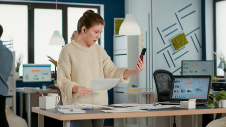 Woman using smartphone in video call conference talking about sales holding paper with charts in...