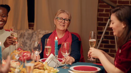 Happy senior woman sitting at table with relatives while enjoying Christmas dinner smiling at camera. Joyful elder person celebrating winter feast with close family members.の写真素材