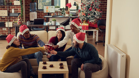 Happy coworkers making toast with wine glasses to celebrate christmas holiday event in office decorated with xmas tree. Festive people with santa hat sayung cheers at winter season festivity.の写真素材