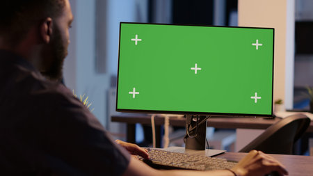 African american man looking at computer with greenscreen, analyzing isolated display on desktop monitor. Using blank copyspace template with mockup screen and chroma key background.の写真素材