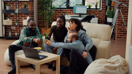 Diverse friends watching funny video on laptop, clinking bottles of beer and enjoying show online on...