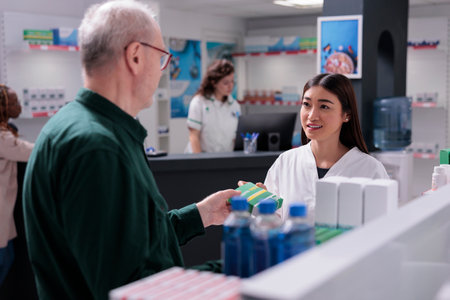 Senior client asking for specialist advice during medication shopping in drugstore. Drugstore worker selling pharmaceutical pills to cure client disease. Pharmacy full with supplements, vitaminの写真素材