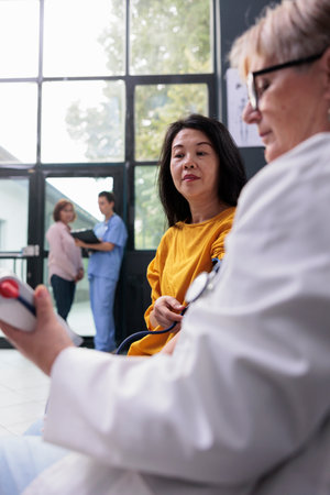 Senior doctor measuring hypertension and blood pressure to do heart exam with tonometer in hospital waiting area. Medic consulting asian patient with cardiology instrument, medicine supportの写真素材