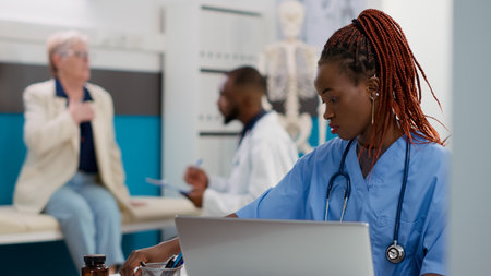 African american nurse working on appointments in cabinet, using laptop to plan checkup visits to help patients at healthcare facility. Medical assistant with stethoscope planning consultations.の写真素材