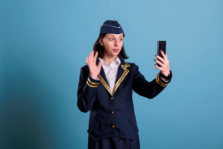 Stewardess talking on smartphone online videocall, having remote conversation, making hi sign. Flight attendant in aviation uniform answering online teleconference call on telephoneの写真素材