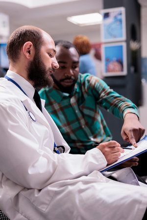 Caucasian general practitioner showing prescription to young african american sick man at hospital reception desk. Male patient giving information about medical condition to doctor in sanatorium.の写真素材