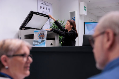 Secretary using copy printer at reception counter to make report papers before checkup consultations. Woman receptionist working on healthcare appointments with forms at registration desk.の写真素材