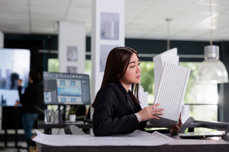 Architect holding building 3d printed model, discussing blueprints on videocall in architecture office. Asian employee talking about architectural design on online meeting in coworking spaceの写真素材