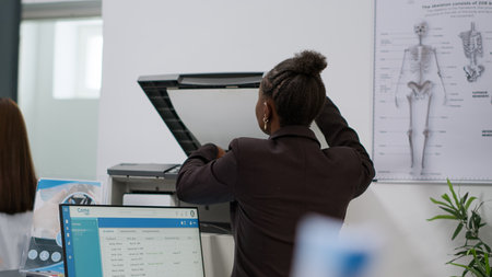 African american woman working at reception desk in health center, using checkup reports with appointments on computer and papers. Receptionist doing registration work for patients with consultations.の写真素材