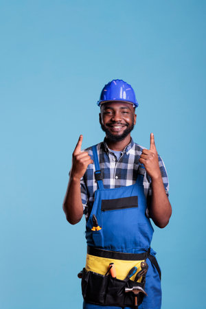 African american construction worker wearing work clothes and hard hat pointing upward with both...