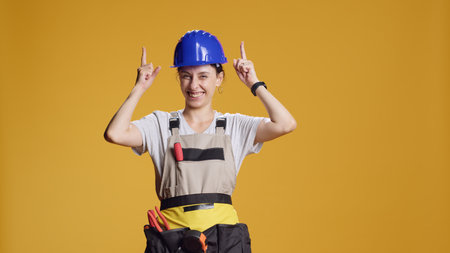 Powerful handywoman pointing up to advertise upwards direction and work on construction building. Contractor indicating above head to show advertisement in studio, engineering uniform.の写真素材
