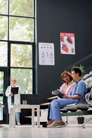 Medical asisstant consulting senior patient in hospital waiting area, filling report papers explaining healthcare treatment. Nurse doing checkup examination with asian woman in lobby.の写真素材