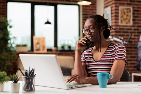 Freelancer talking on smartphone with client, working on laptop, in home office. Busy remote worker answering phone call, employee in glasses having conversation on telephoneの写真素材