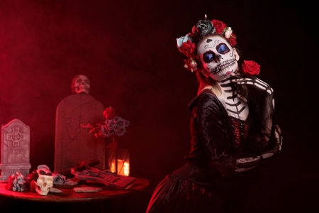 La cavalera catrina wearing holy costume and flowers crown, holding roses and acting spooky in studio. Santa muerte model with skull make up celebrating traditional dios de los muertos.の写真素材