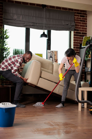 African american man lifts sofa while woman dusts underneath with mop, roommates doing spring...