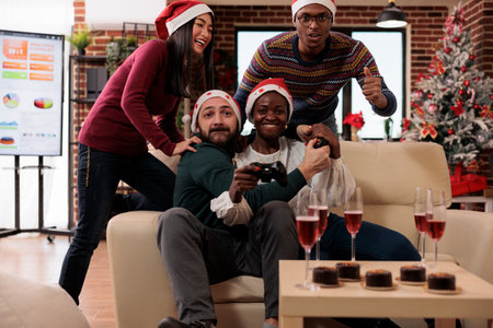 Happy coworkers having fun with video games at event in festive office decorated with xmas tree and...