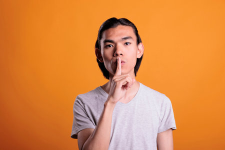 Young adult asian man showing quiet gesture, making tsss sign, silence concept. Confident teenager loking at camera with forefinger on lips, attractive person keeping secret, studio mid shotの写真素材