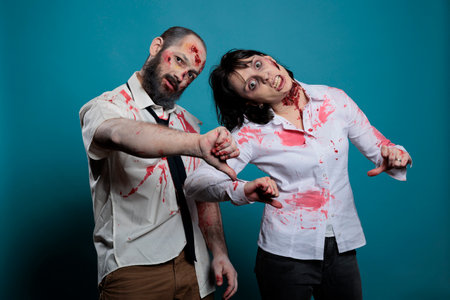 Creepy halloween monsters showing thumbs down on camera, expressing disapproval and dislike in studio. Couple of brain eating zombies with bloody scars and ugly wounds, frightening devils.の写真素材
