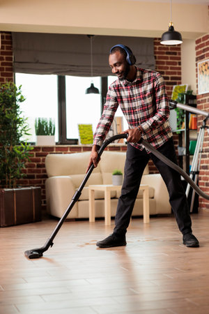 Smiling African american man with wireless headset cleaning floor at home with vacuum cleaner. Guy taking care of household chores, doing spring cleaning in urban apartment.の写真素材