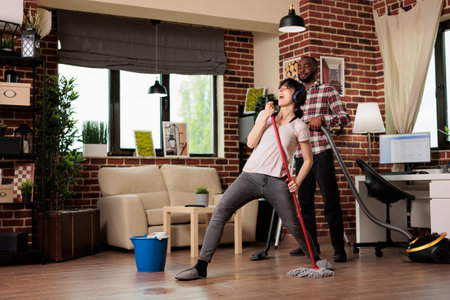 Married multiracial couple cleaning apartment, husband disappointed to be cleaning while wife just having fun singing with mop. Woman joking without helping, husband does chores by himself.の写真素材