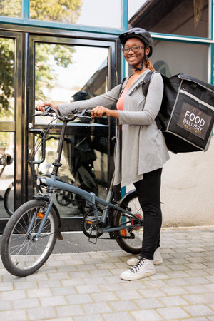 Courier in bicycle helmet waiting for customer outdoors portrait, african american woman standing near office building entrance, looking at camera. Takeaway food delivery serviceの写真素材