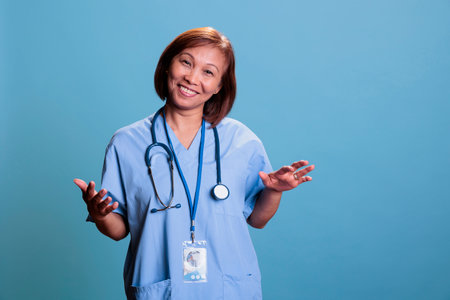 Happy asian nurse wearing stethoscope and blue uniform during checkup visit consultation working at health care treatment. Physician assistant smiling at camera standing in studio with blue backgroundの写真素材