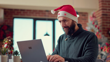 Young adult brainstorming ideas to create report on laptop, wearing santa hat in office with christmas tree. Festive man working on computer with research data during holiday season. Handheld shot.の写真素材
