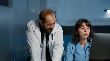 Clinical staff looking at computer analyzing medical expertise while discussing medication treatment to help cure patient disease. Physician and nurse working over hours in hospital office.の写真素材