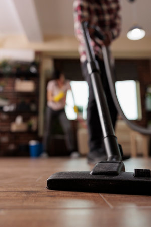 Man vacuuming apartment floor while woman in background dusts shelves. Modern couple doing household chores, finishing spring cleaning and disinfecting spaces, out of focus image.の写真素材