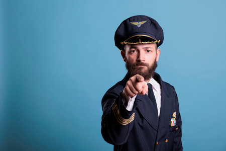 Serious airplane aviator pointing at camera with finger, aviation academy pilot training recruitment. Confident finger wearing air crew uniform and hat front view portraitの写真素材