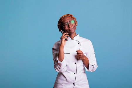 African american chef with confused expression on phone call. Bewildered afro female kitchen worker on cell phone conference. Curly haired girl cooker on strange audio call.の写真素材