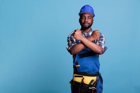 Builder making gesture of declining crossed arms while looking at camera. African american construction worker in work uniform giving declining hands symbol while standing on blue background.の写真素材