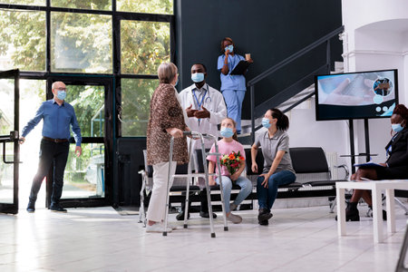 Eldely woman with walking frame discussing medical diagnosis with medic during checkup visit in hospital waiting area. People wearing protective face mask to prevent infection with coronavirusの写真素材