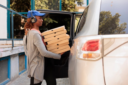 Office lunch delivery service worker delivering pizza by car, side view. African american courier in headphones holding fastfood boxes stack, standing near company building outdoorsの写真素材