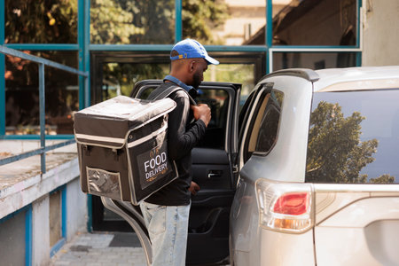 Young man delivering office food order by car. Takeaway lunch delivery service worker holding thermal backpack with meal, standing near vehicle outdoors, side view medium shotの写真素材
