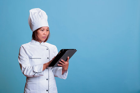 Serious chef with apron and white uniform holding tablet computer browsing culinary recipe on gastronomy site. Cheerful senior woman cooking dinner meal using healthy ingredient, food preparationの写真素材