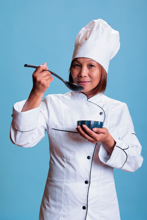 Confident positive cook holding kitchen bowl tasting prepared food meal in studio with blue background. Cheerful chef cooking dinner dish using healthy ingredients, gastronomy industryの写真素材