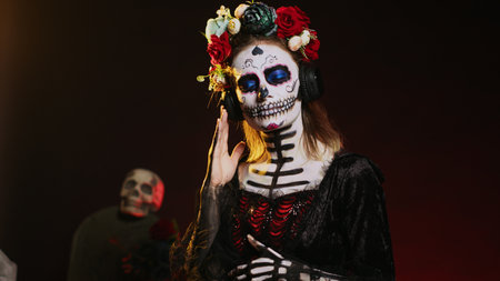 Lady of death listening to music on headphones, wearing skull make up and santa muerte costume on day of the dead. Enjoying fun song on audio headset, celebrating mexican tradition. Handheld shot.の写真素材