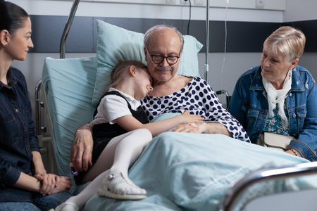 Elderly man in geriatric clinic bedroom hugging little girl. Senior patient relatives visiting medical tower recovery room. Granddaughter visiting sick grandfather at sanatorium.の写真素材