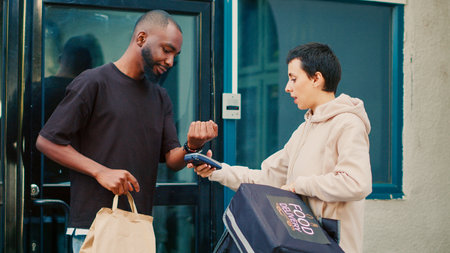 African american man using smartwatch to make nfc payment on pos terminal, paying for restaurant takeaway meal. Female delivery courier delivering fastfood package at front door. Handheld shot.の写真素材