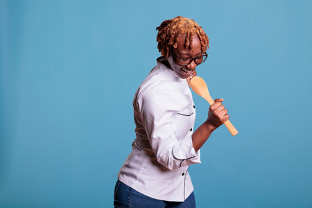 Cheerful woman holds a spatula in hand simulating microphone, dances and sings with excitement. Energetic female chef wears white work jacket in studio shot against blue background.の写真素材
