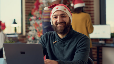 Portrait of festive man sitting at desk to work during holiday season in office with christmas tree and decorations. Employee wearing santa hat to give presents on winter festivity.の写真素材