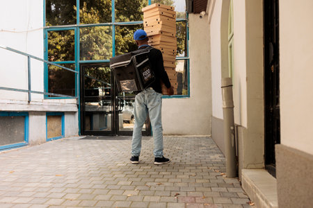 Courier carrying huge pizza boxes pile back view, deliveryman holding food order, standing near office building. Lunch delivery service african american worker delivering fastfoodの写真素材