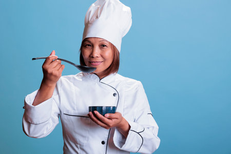 Cheerful cook with restaurant uniform holding bowl with culinary meal while tasting food before start eating, cooking healthy recipe. Professional asian chef preparing dinner dish, gastronomy industryの写真素材