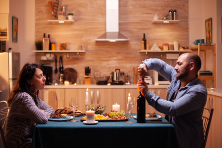Husband opens wine bottle while celebrating relationship with wife in kitchen. Happy couple talking, sitting at table in kitchen, enjoying the meal, celebrating their anniversary at home.の写真素材