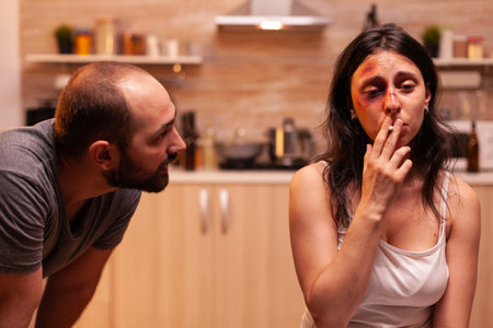 Woman suffering after husband beating and smoking sitting on chair in kitchen. Violent aggressive man abusing injuring terrified helpless, vulnerable, afraid, and panicked woman.の写真素材