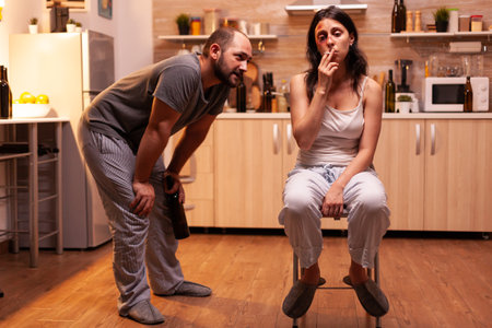 Victim of domestic violence smoking sitting on chair while drunk husband looking at her. Violent aggressive man abusing injuring terrified helpless, vulnerable, afraid, beaten and panicked woman.の写真素材