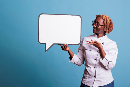 Woman smiling kitchen staff member pointing to blank dialogue cloud with copy space. Cook holding dialogue bubble with message frame looking at camera, studio shot on blue background.の写真素材