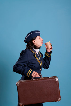 Flight attendant in professional airline uniform carrying suitcase, drinking coffee to go. Stewardess with luggage in airport, air hostess with baggage holding tea paper cup, side viewの写真素材