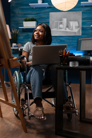Female sketch artist wheelchair user using laptop computer to take drawing classes remotely at home...